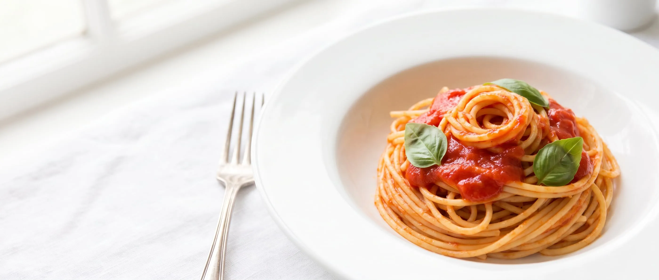 A white plate of spaghetti with tomato sauce and basil leaves in soft natural light